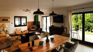 Een ruime woonkamer met een houten eettafel, comfortabele zithoek, een open haard en een groot raam met uitzicht op groen.
