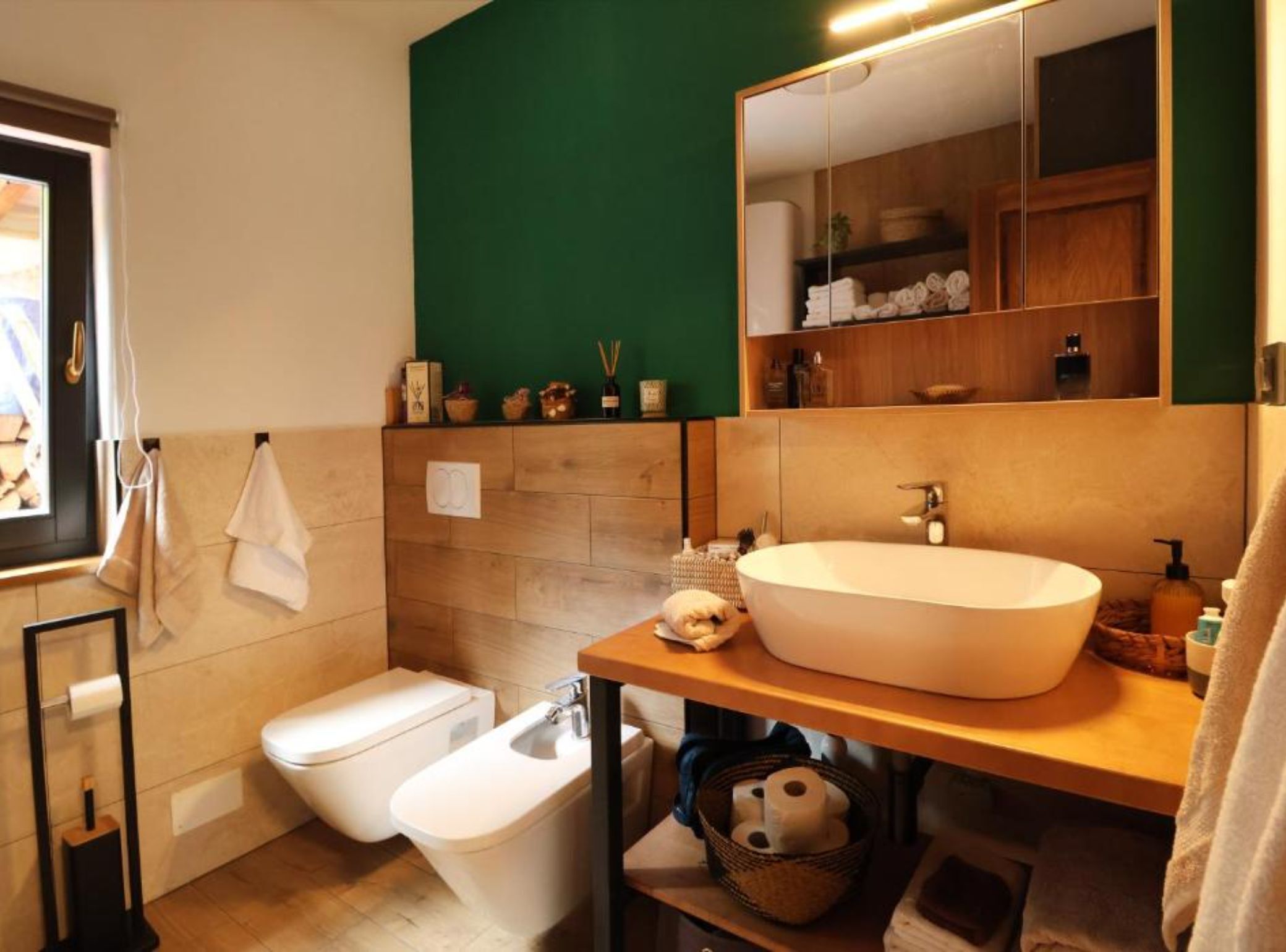 Een moderne badkamer met een vrijstaand bad, een toilet en een douchetoilet. Boven de wastafel hangt een spiegel, en er staan diverse toiletartikelen op het aanrecht.