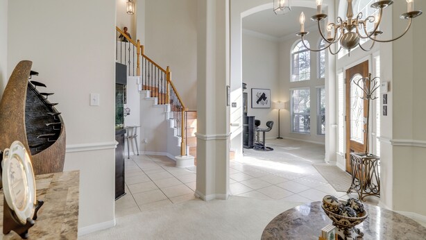 A spacious foyer with a grand staircase, a chandelier, and a large decorative bowl on a marble countertop. - Lake-view Home w/ Pool Access in Legends Ranch! (Spring)