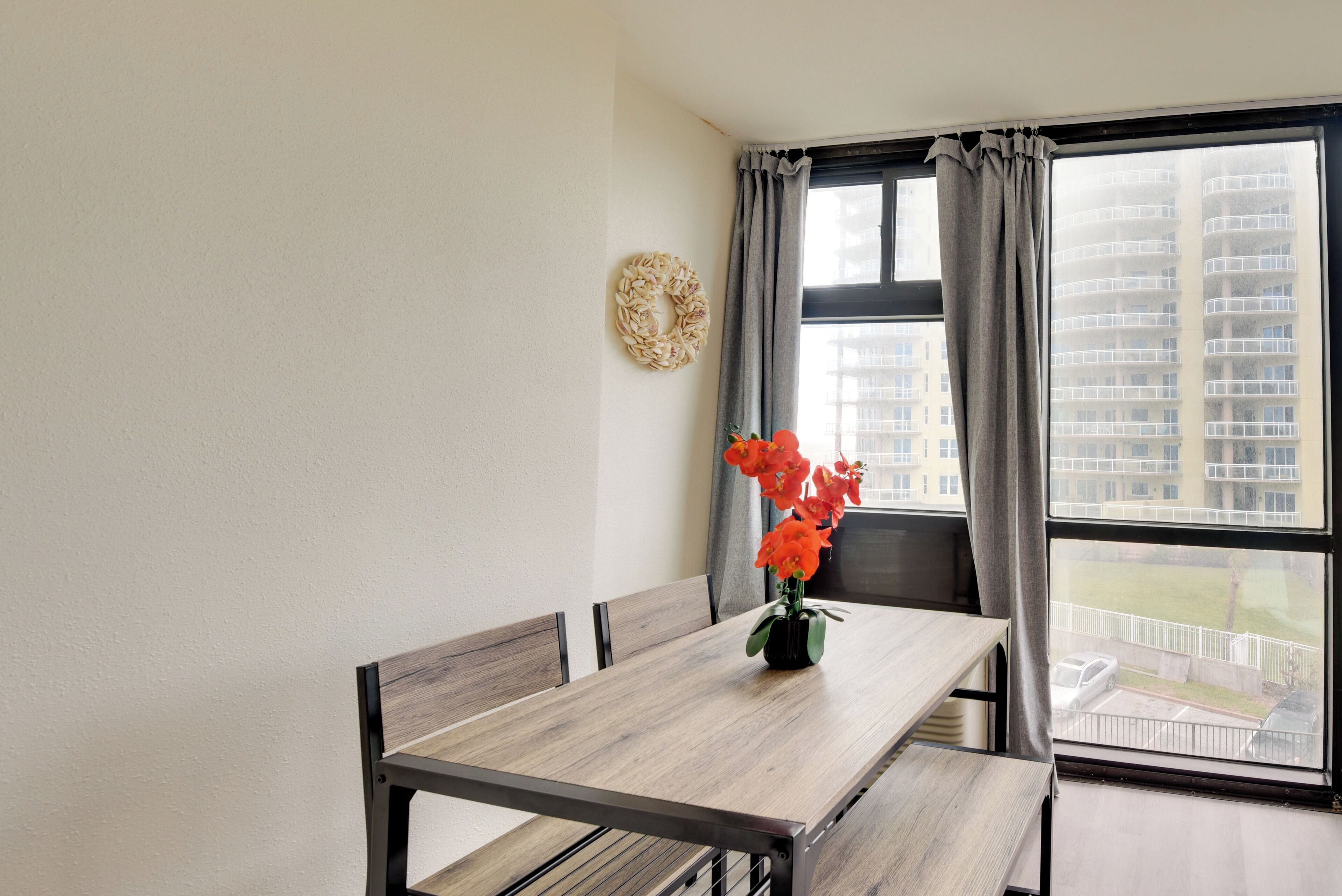 A dining area with a wooden table and chairs, a vase with flowers, and a view of a building through a window with curtains.