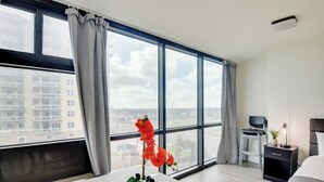 A modern hotel room with large windows, a wooden table with a vase of red flowers, a black nightstand with a lamp, and a bed with white bedding. - Oceanfront Studio Amazing Beach Views Pool (Daytona Beach Shores)