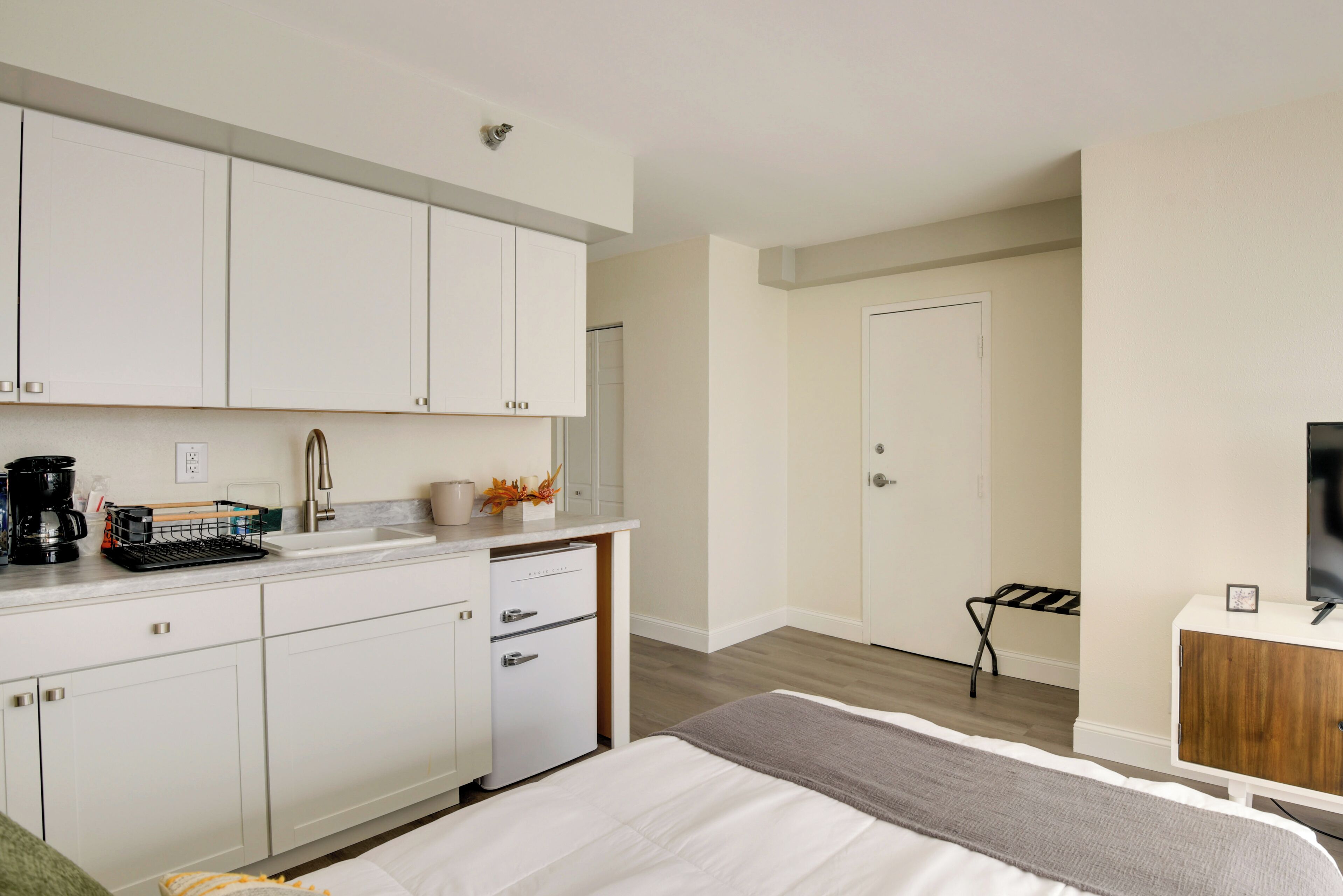 A modern hotel room with a kitchenette, a bed with a gray blanket, a TV on a wooden cabinet, and a door leading to a bathroom.