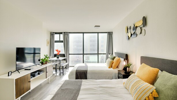 A modern hotel room with a large bed, a desk, a TV, and a view of the city through large windows. - Oceanfront Studio Amazing Beach Views Pool (Daytona Beach Shores)
