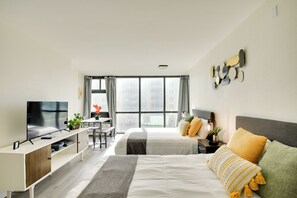 Ein modernes Hotelzimmer mit einem großen Bett, einem Schreibtisch, einem Fernseher und Blick auf die Stadt durch große Fenster.