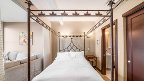 A four-poster bed with a metal frame and white bedding, a sofa with cushions, a bedside table with a lamp, and a wooden door.