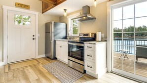 Una cocina moderna con refrigerador, horno y estufa de acero inoxidable, vigas de techo de madera y vista a un cuerpo de agua a través de un ventanal grande.