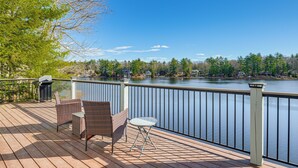 Terraza de madera con sillas de mimbre y una mesita pequeña con vistas a un lago, con barandilla y una parrilla.