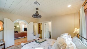 Ein Schlafzimmer mit einem Bett, Deckenventilator, einem fenster mit Vorhängen und einem Flur, der zu einem Badezimmer führt.