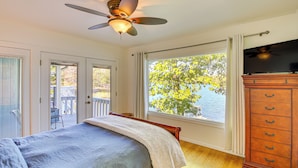 A bedroom with a bed, a ceiling fan, a dresser, and a large window with a view of a dock and trees.