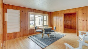 Une pièce aux murs en bois, une table ronde entourée de chaises, une grande fenêtre et un banc blanc.