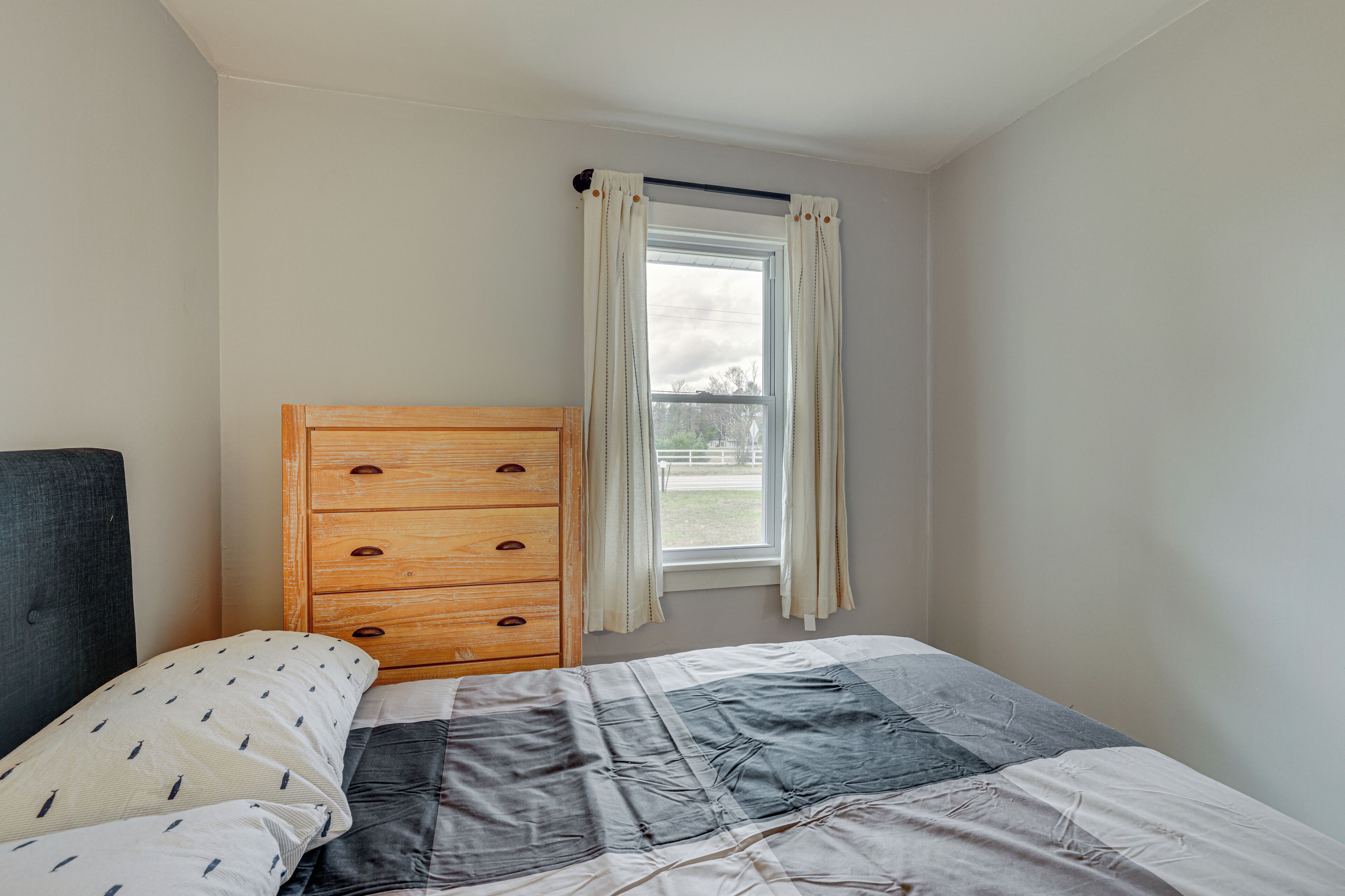 A bedroom with a bed, a wooden dresser, a window with curtains, and a view of the outside.