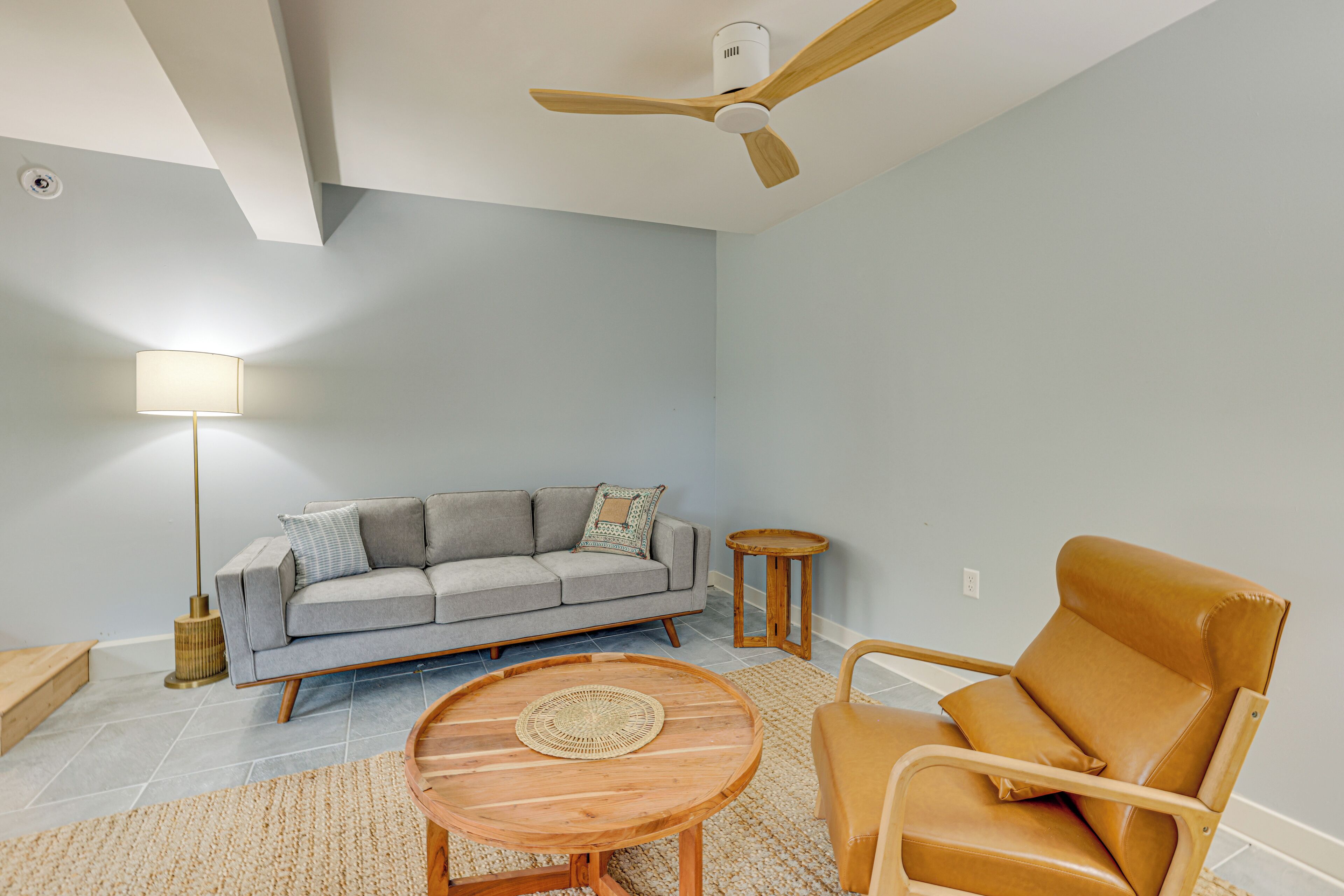 A modern living room with a sofa, a round wooden coffee table, a leather armchair, a floor lamp, and a ceiling fan.