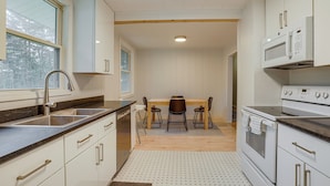 Une cuisine moderne avec des meubles blancs, un évier en acier inoxydable, une table à manger en bois avec quatre chaises et un four encastré.