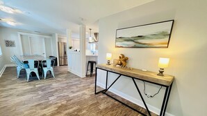 Una sala moderna con piso de madera, un comedor con sillas azul claro y una cocina con gabinetes blancos, además de un aparador con dos lámparas.