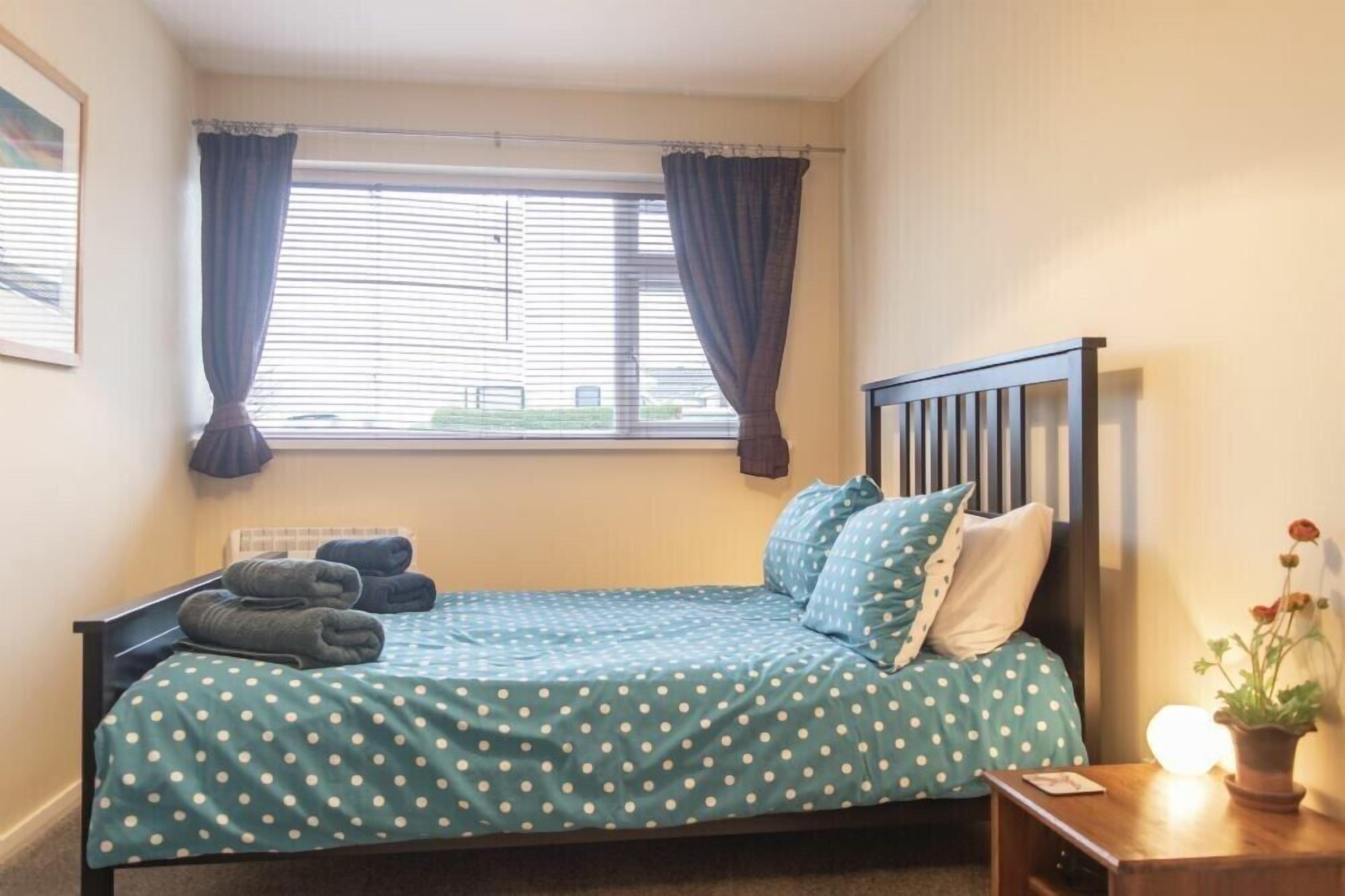 A neatly made bed with a polka dot bedspread, a wooden headboard, and a bedside table with a lamp and a potted plant.