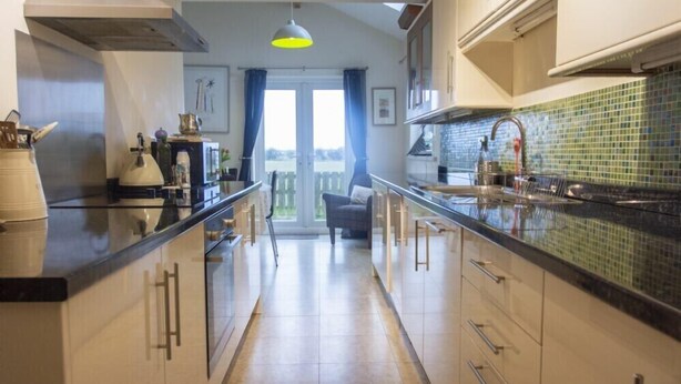 A modern kitchen with a large island, a view to the outside, and a dining area with a chair. - Our Story Cottage (Alnwick)