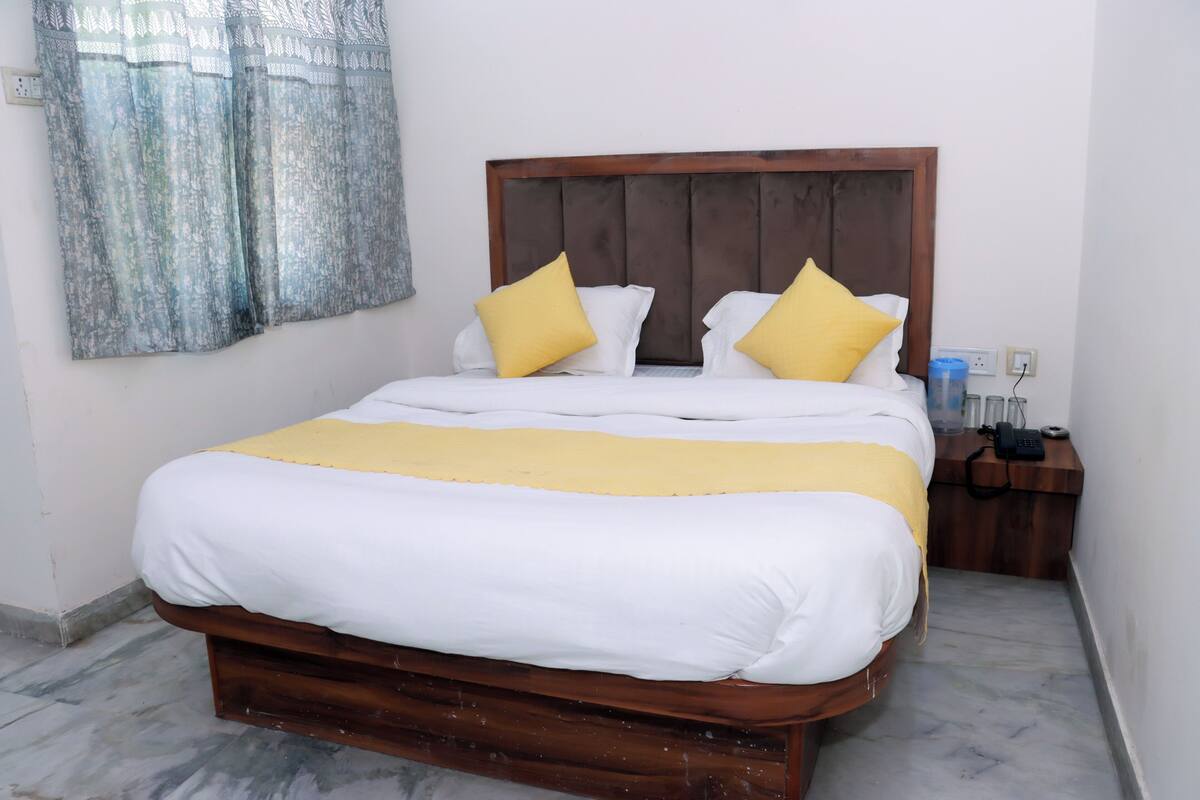A neatly made bed with white and yellow bedding, a wooden headboard, and a bedside table with a phone and a water bottle.