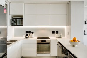 A modern kitchen with white cabinetry, a built-in oven, a dishwasher, and a stainless steel refrigerator. - Cape Town Deluxe City Apartment Hafen Views (Cape Town)
