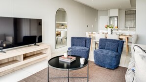 A modern living room with a flat-screen TV, a coffee table, and two blue armchairs. - Cape Town Deluxe City Apartment Hafen Views (Cape Town)