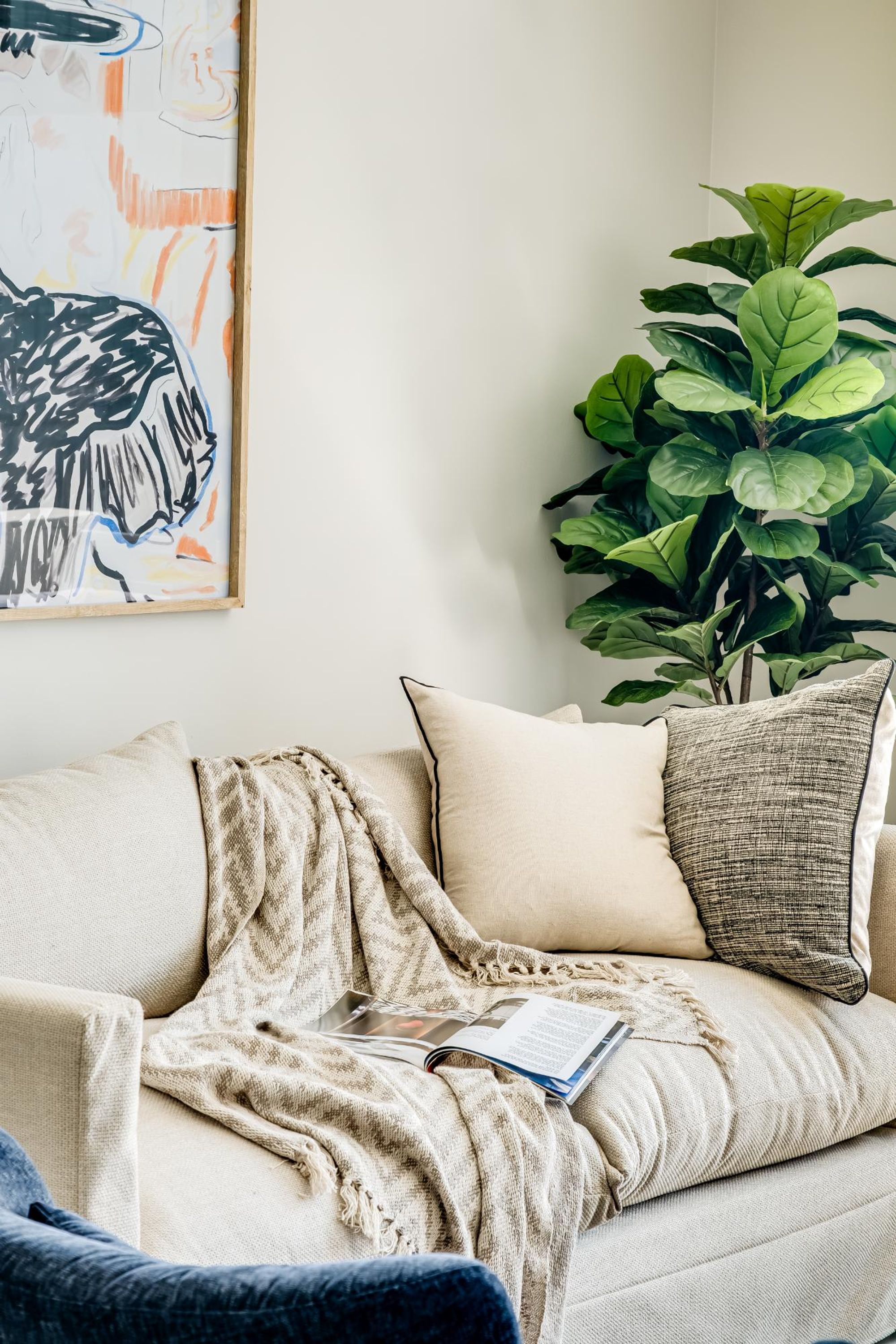 Een gezellige woonkamer met een bank, kussens, een plaid, een plant en een abstract schilderij in een lijst.
