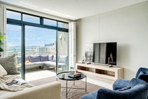A modern living room with a large TV, comfortable seating, and a view of the city through large windows. - Cape Town Deluxe City Apartment Hafen Views (Cape Town)