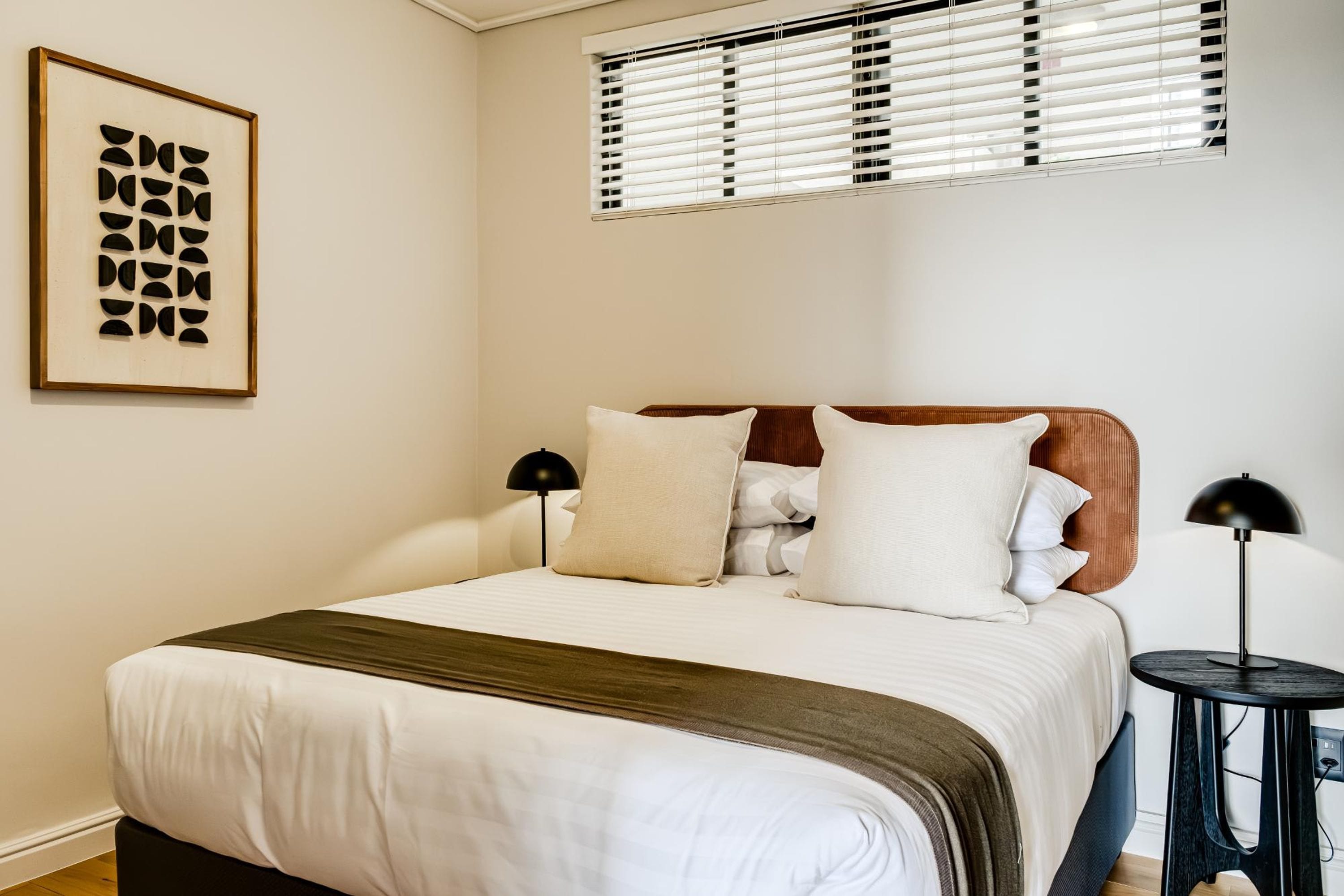 A neatly made bed with a wooden headboard, white bedding, and a brown throw blanket. There is a framed abstract art piece on the wall and a window with blinds above the bed.