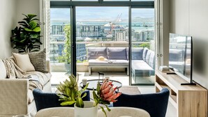 Una sala moderna con un sofá, una mesa de centro y un televisor. Un ventanal ofrece vistas a un paisaje urbano y al agua.