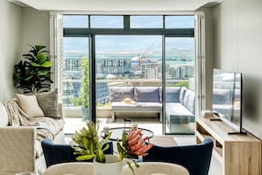 A modern living room with a sofa, a coffee table, and a TV. A large window offers a view of a cityscape and water. - Cape Town Deluxe City Apartment Hafen Views (Cape Town)
