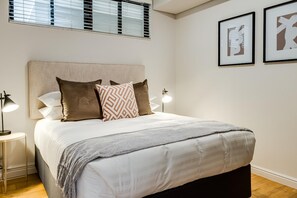 Una cama bien hecha con un cabecero, una mesita de noche con una lámpara y dos cuadros enmarcados en la pared.