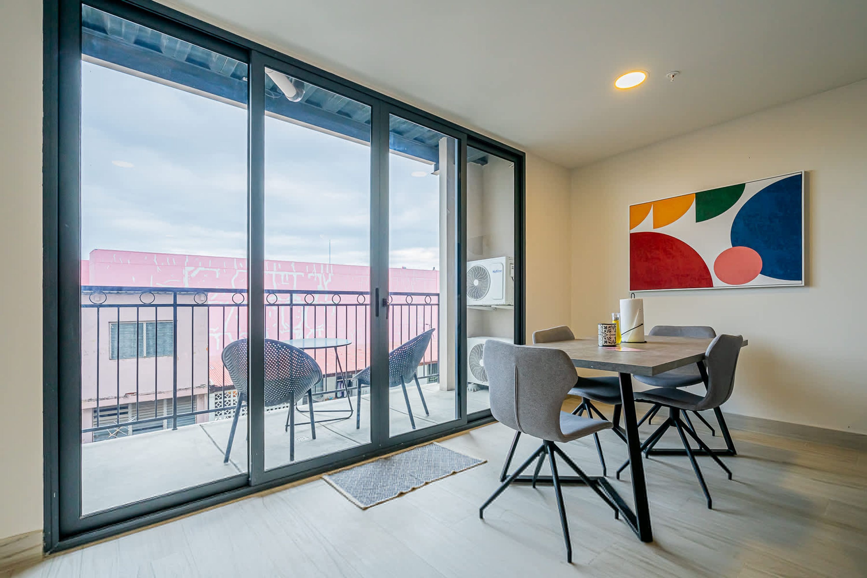 A dining area with a table and chairs, large windows, and a colorful abstract painting on the wall.