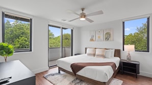 Une chambre à coucher avec un lit, une table de chevet, un bureau et un ventilateur de plafond. De grandes fenêtres offrent une vue sur les arbres à l’extérieur.