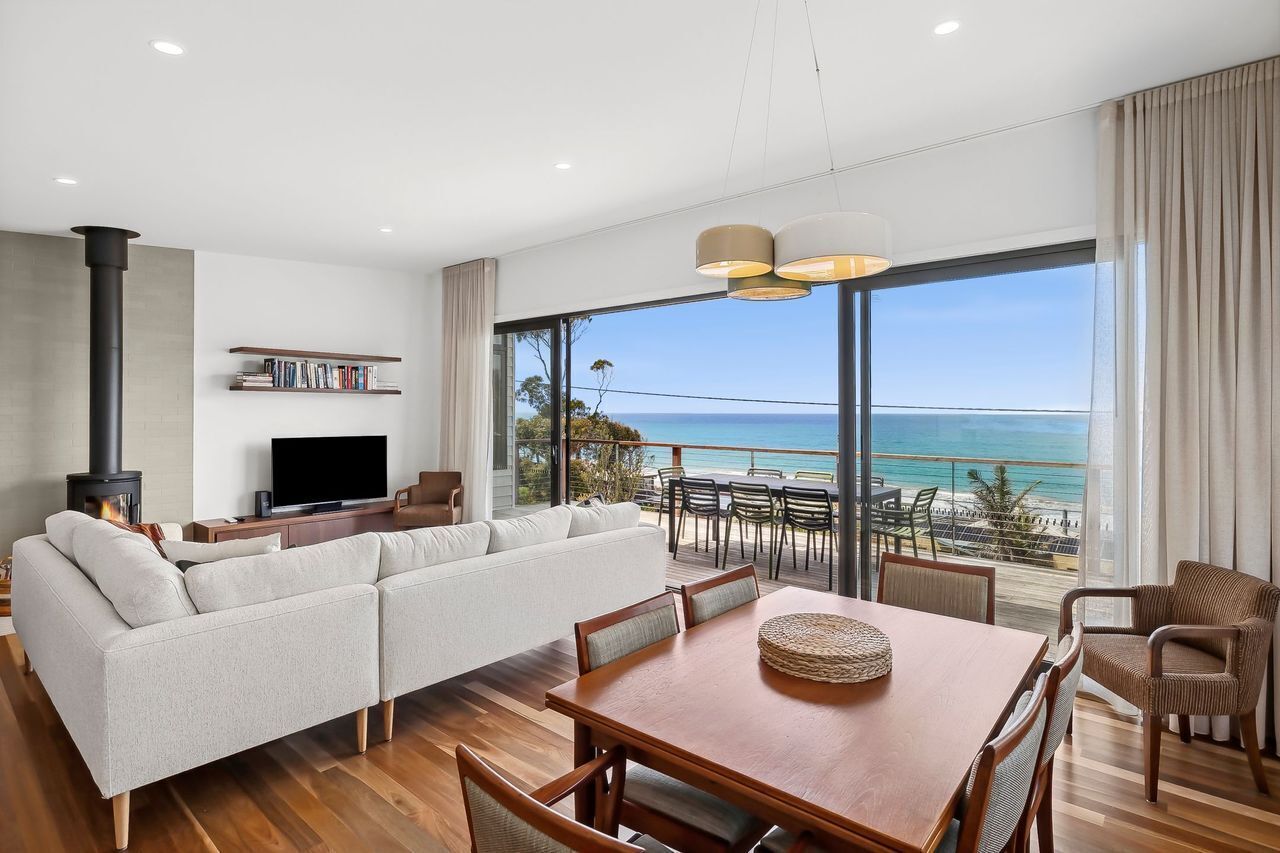 Een moderne woonkamer met een grote bank, een eettafel en een televisie. Er is een open haard, een boekenkast en een balkon met uitzicht op de oceaan.