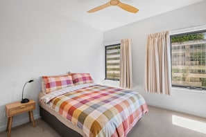 A bedroom with a bed, a nightstand, a ceiling fan, and two windows with curtains.