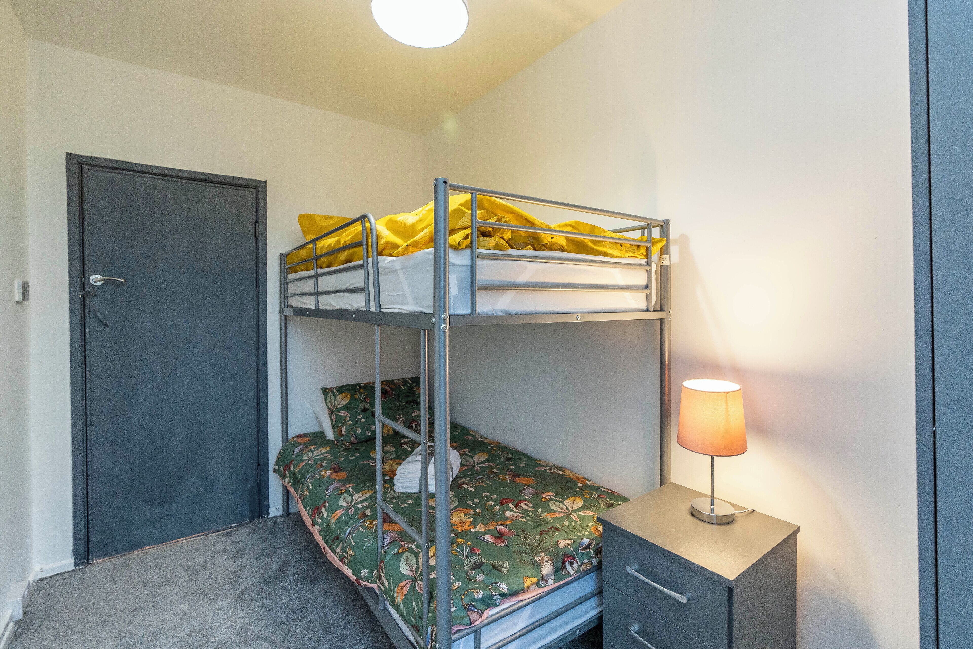 Una cama litera con una colcha floral verde, una mesita de noche con una lámpara y una puerta.