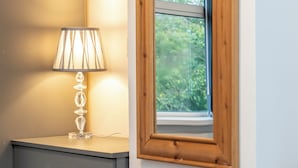 Une table de chevet avec une lampe, une fenêtre à cadre en bois et un miroir fixé au mur.