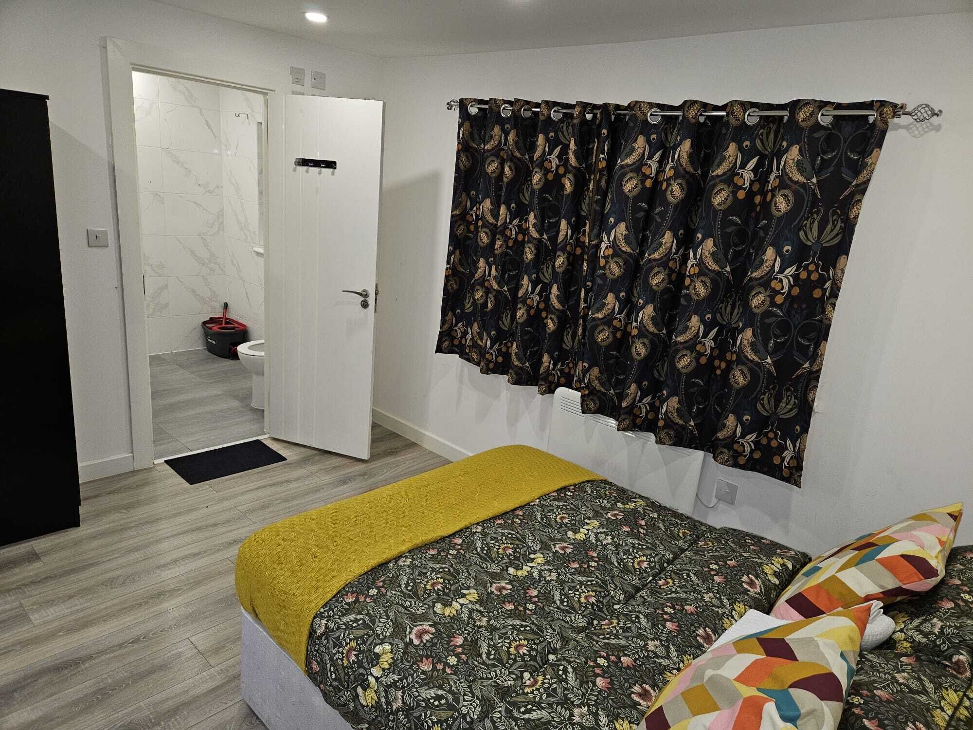 A bedroom with a bed, a yellow throw, a patterned bedspread, and a curtain with a bird pattern.