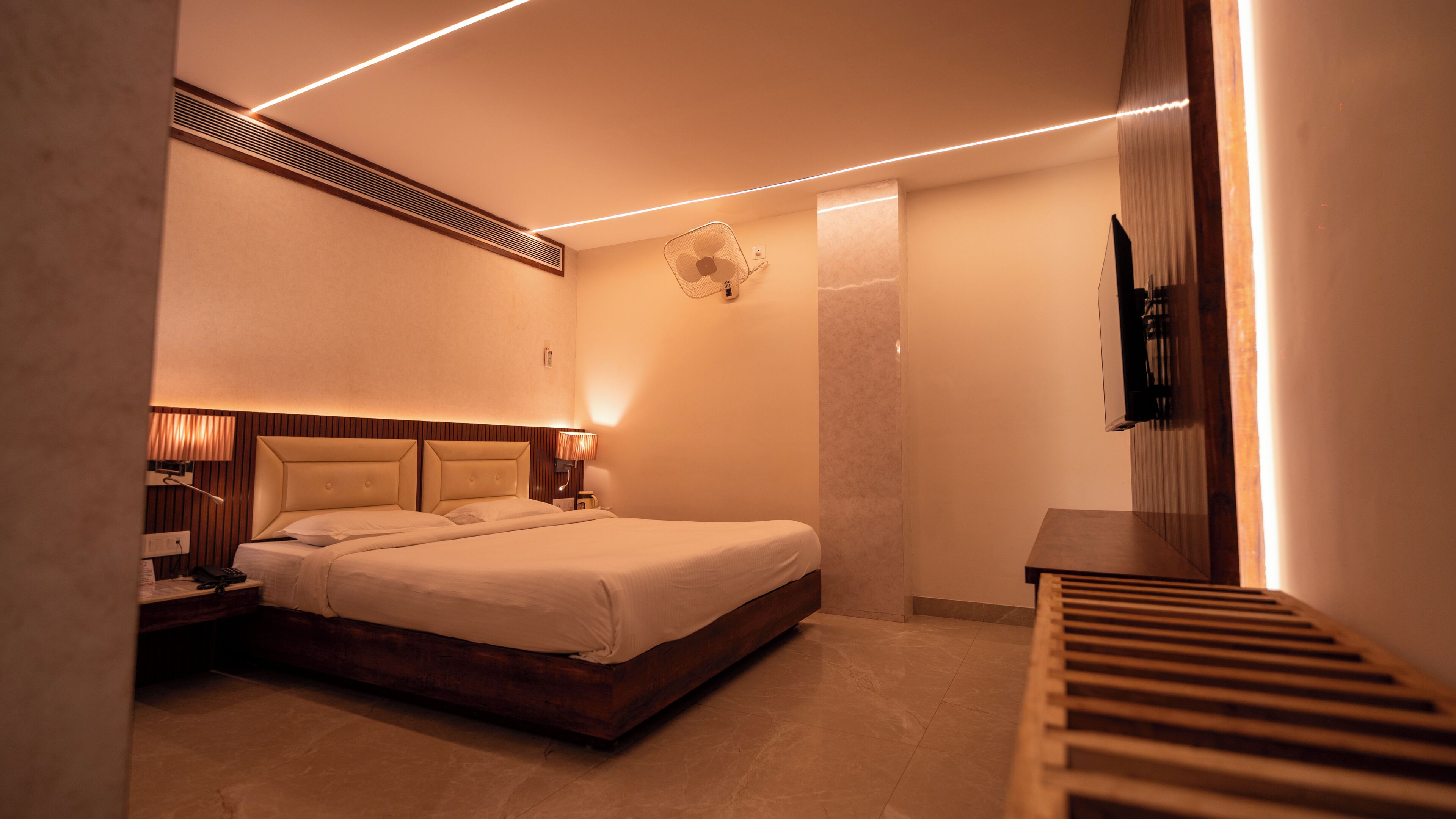 A modern hotel room with a large bed, bedside lamps, a wall-mounted fan, and a TV on a wooden stand.