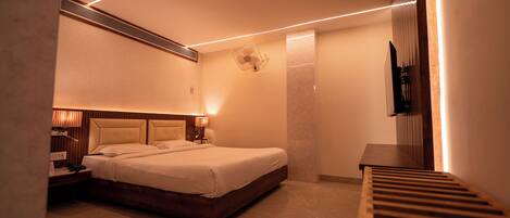 A modern hotel room with a large bed, bedside lamps, a wall-mounted fan, and a TV on a wooden stand.