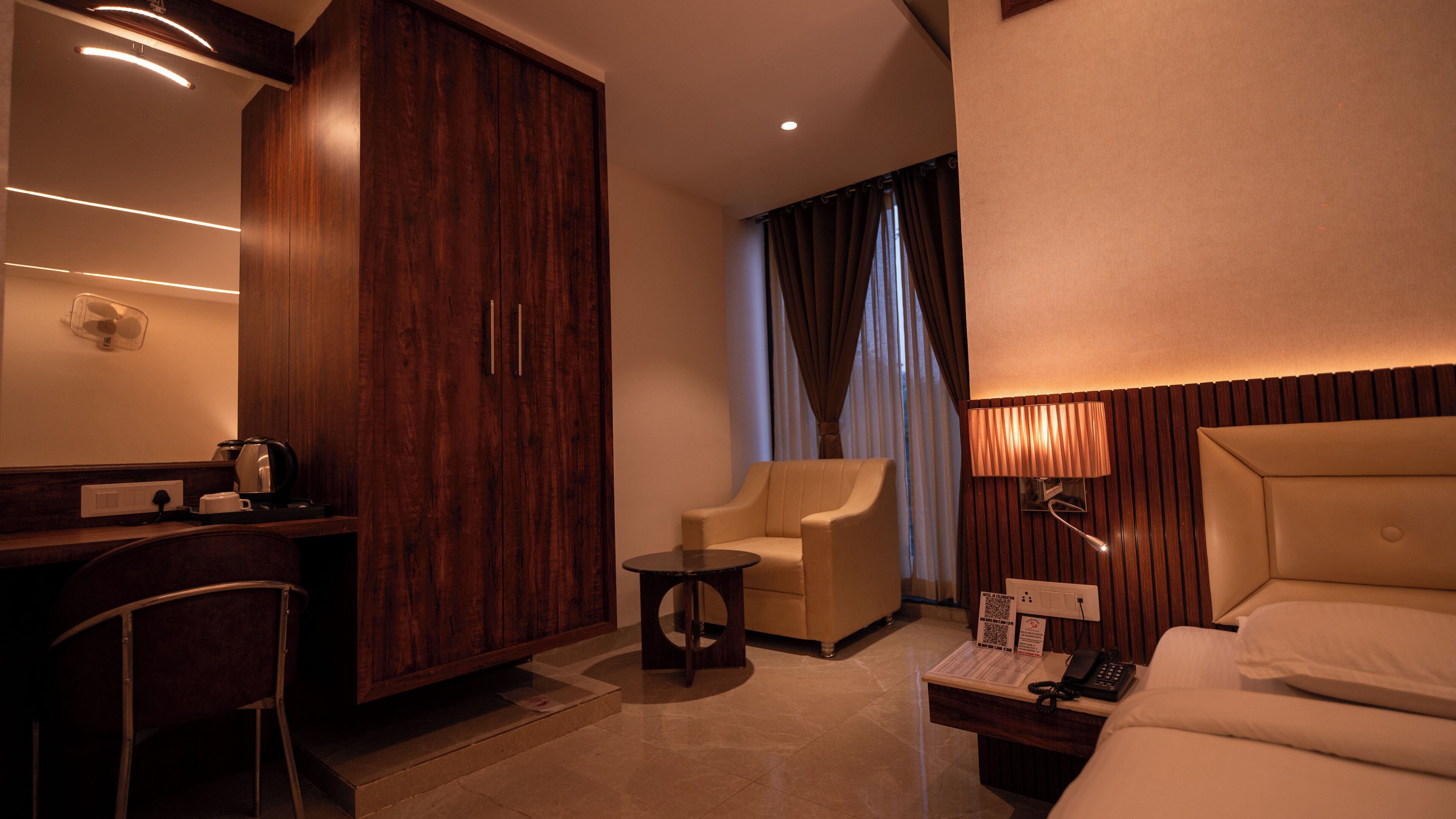 Habitación de hotel con armario de madera, una pequeña mesa redonda, sofá, lámpara de noche, cama y escritorio con hervidor y ventilador.