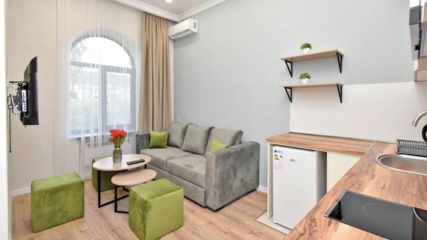 A modern living room with a grey sofa, green ottomans, a small round table with a vase of flowers, and a compact kitchen area with a fridge and wooden countertop. - Apartment on Khanjyan street (Yerevan)