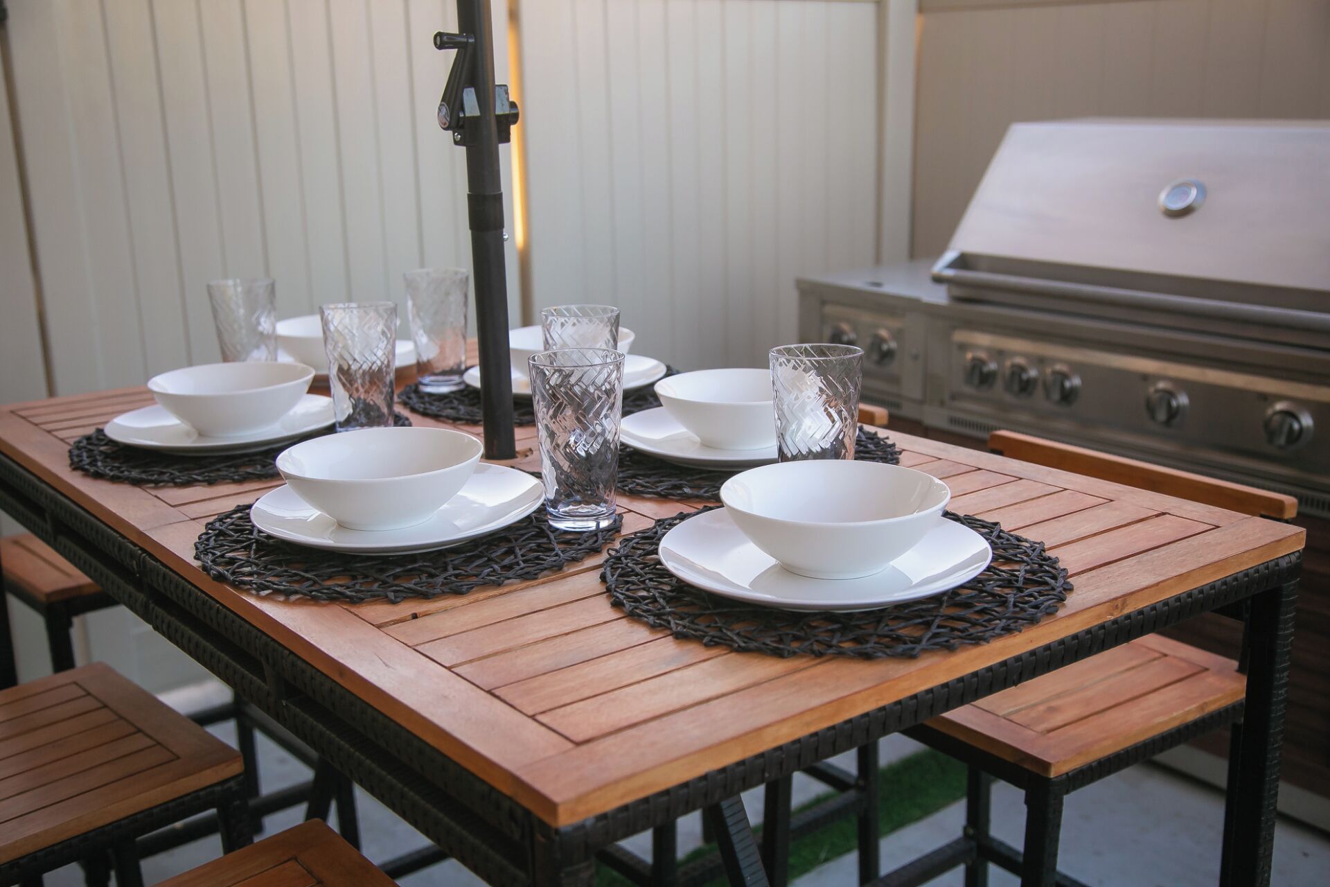 A wooden dining table set with white bowls and glasses, a grill in the background, and a black metal stand in the center.
