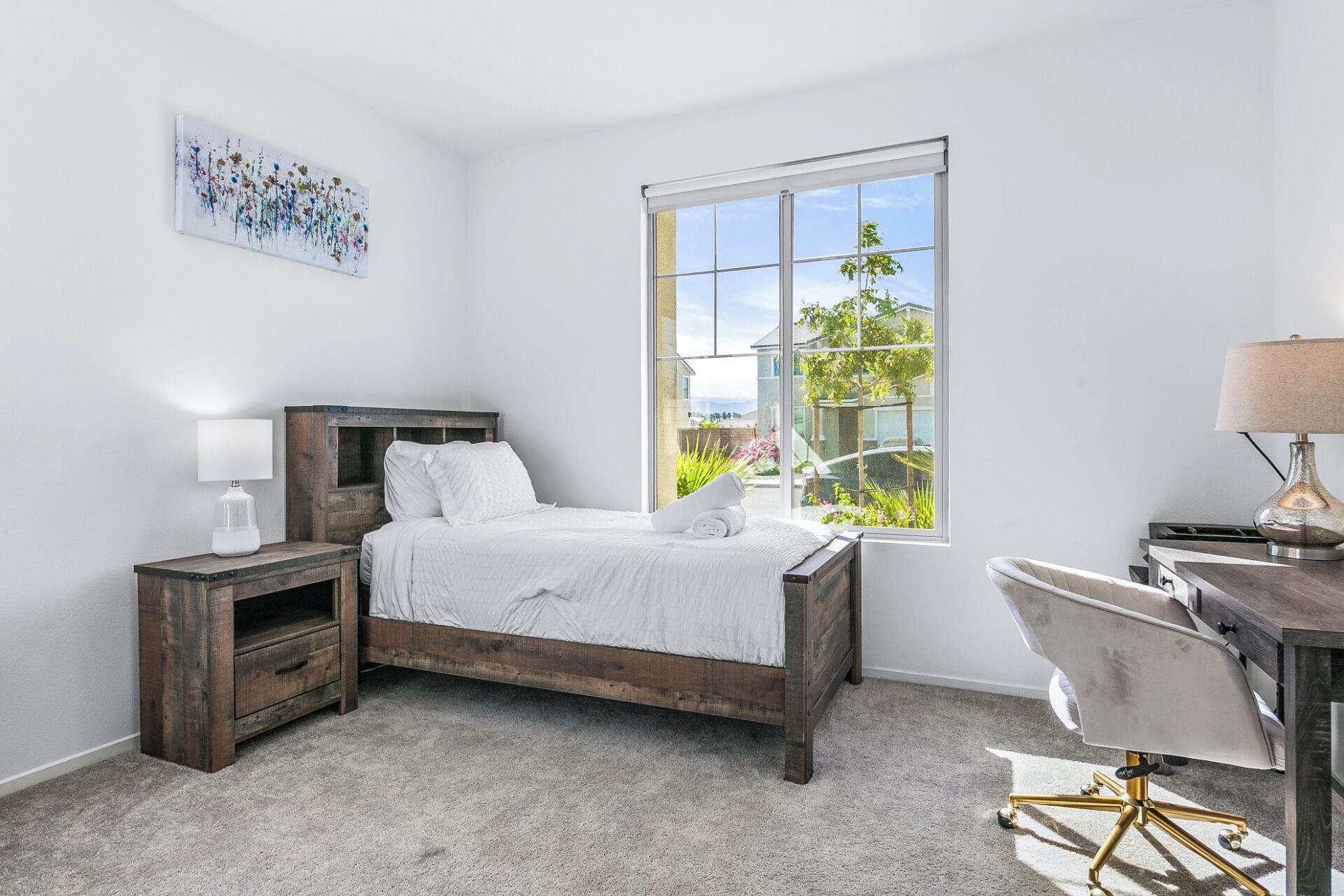 A bedroom with a wooden bed, a nightstand, a desk with a chair, a window with a view of greenery, and a painting on the wall.