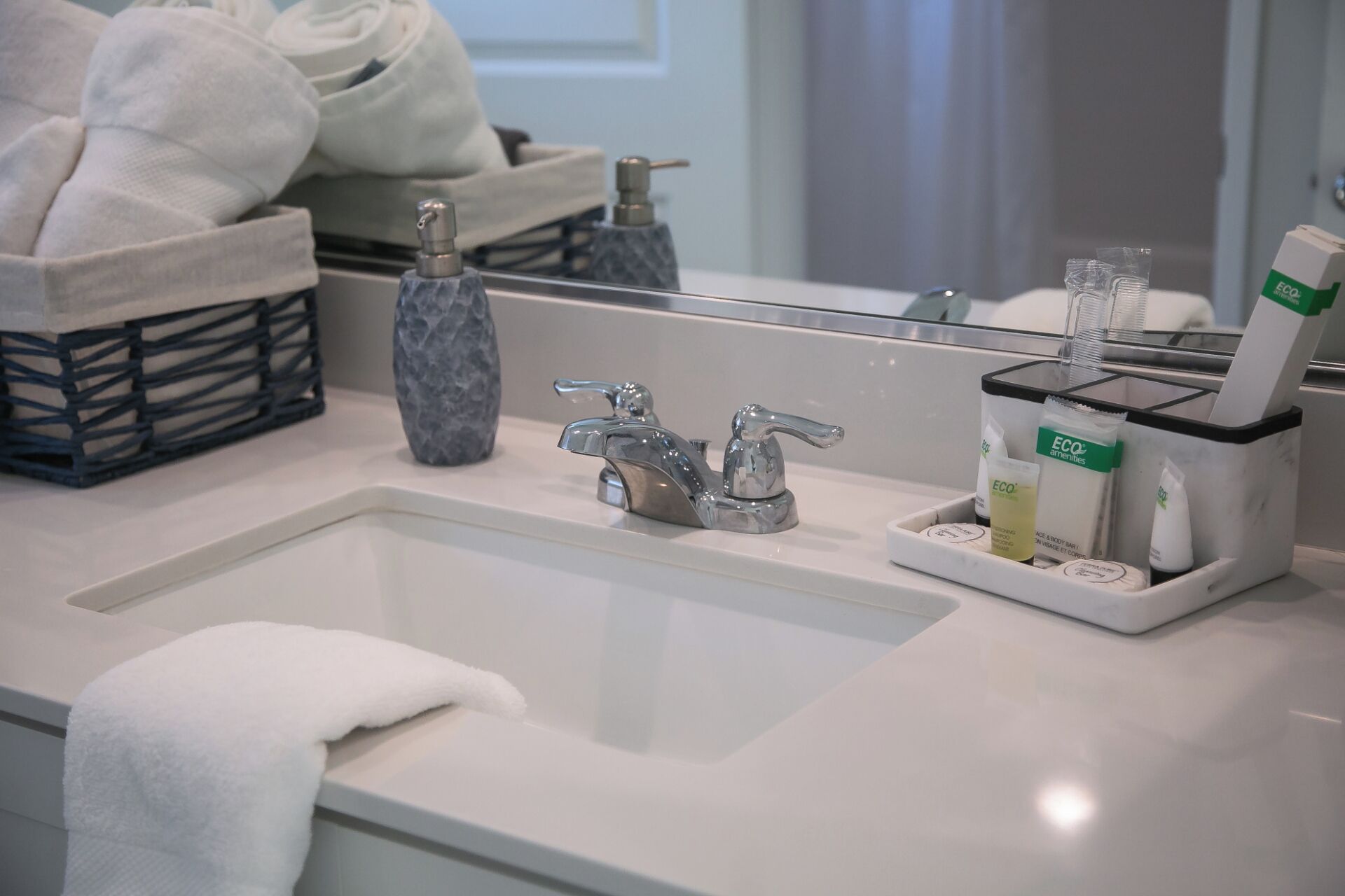 A bathroom with a white sink, a mirror, and a towel rack. There are toiletries on the sink and a bottle of hand soap.