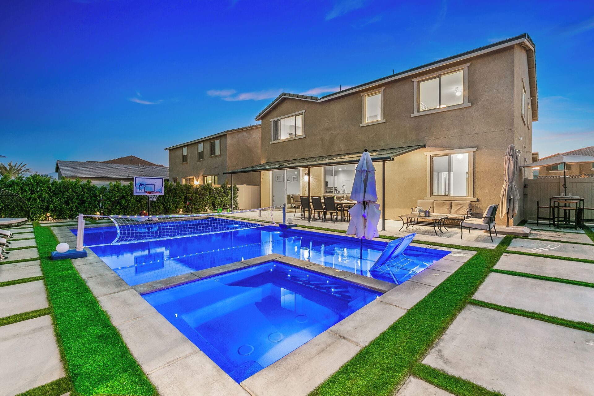 A rectangular swimming pool with a netted area, surrounded by a patio with chairs and a table, and a house in the background.