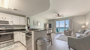Una cocina moderna con armarios blancos, un microondas y una isla grande. Hay un área de comedor con una silla y un sofá. La habitación tiene un balcón con vista al océano.