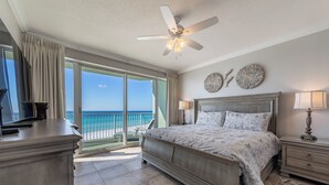 A bedroom with a large bed, a ceiling fan, a TV, and a view of the ocean.