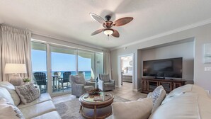 A spacious living room with a large flat-screen TV, comfortable seating, a ceiling fan, and a balcony with outdoor furniture.