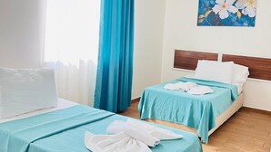 A hotel room with two beds, each with a blue bedspread and white pillows, a wooden headboard, a painting of flowers on the wall, and an air conditioning unit on the wall. - ŞİRİN APARTROOMS (Denizli)