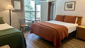 A hotel room with a bed, a desk, and a chair. There is a window with a view of trees and a door leading to another room. - Mountainside Inn 115/117 2 Bedroom Hotel Room (Telluride)