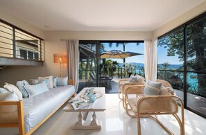 A modern living room with a sofa, coffee table, and wicker chairs, overlooking a pool and greenery.